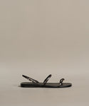 The Romy Sandal is a black minimalist design with thin leather straps and a small gold buckle, handmade in Tuscany and shown in profile against a plain beige background.