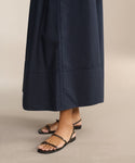 A person in a long, dark navy skirt wears Romy Sandals, handcrafted in Tuscany, standing on a beige floor. Only the lower legs and feet are visible.