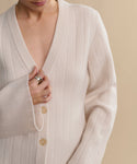 A person wears the Rory Cardigan, a cream ribbed V-neck knit with large buttons, slightly open at the chest. They hold it closed with one hand, showing a silver ring against a neutral beige background.