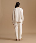 A model viewed from behind wearing a matching cream-colored knit lounge set consisting of a crewneck sweatshirt and jogger pants, paired with tan flats against a solid beige studio backdrop.