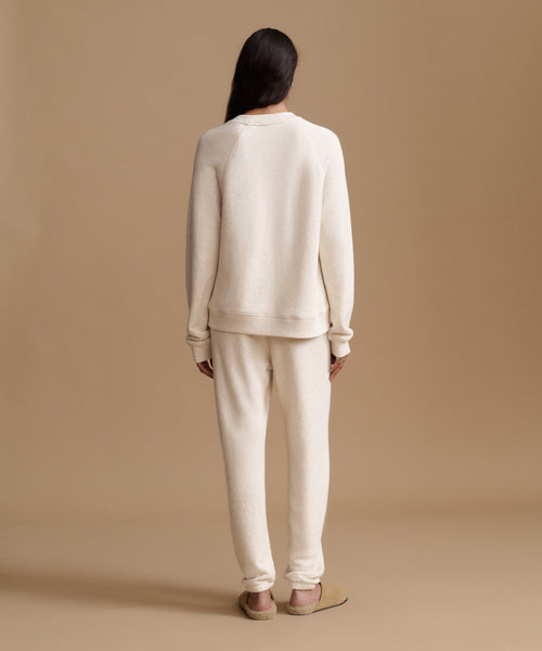 A model viewed from behind wearing a matching cream-colored knit lounge set consisting of a crewneck sweatshirt and jogger pants, paired with tan flats against a solid beige studio backdrop.