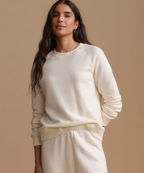 A woman modeling a cream-colored crewneck sweatshirt and matching sweatpants against a solid beige background.