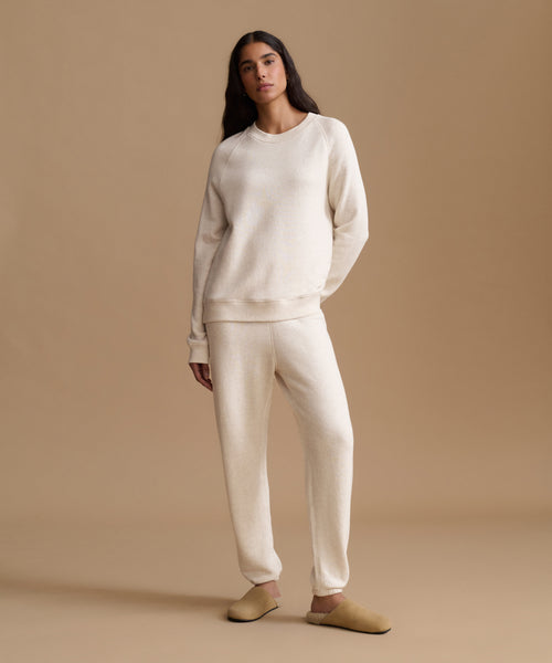 A woman modeling a matching cream-colored lounge set consisting of a crewneck sweatshirt and jogger pants, paired with tan slip-on shoes, against a solid beige studio background.