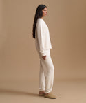 A woman stands in profile wearing a matching cream knit lounge set consisting of a long-sleeve sweatshirt and wide-leg sweatpants, paired with tan slip-on shoes, against a solid beige studio background.