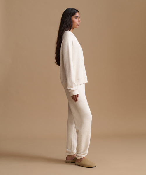 A woman stands in profile wearing a matching cream knit lounge set consisting of a long-sleeve sweatshirt and wide-leg sweatpants, paired with tan slip-on shoes, against a solid beige studio background.