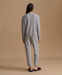 A woman viewed from behind wearing a light gray crewneck sweatshirt and matching jogger pants, paired with black slip-on slippers, against a solid warm beige studio background.