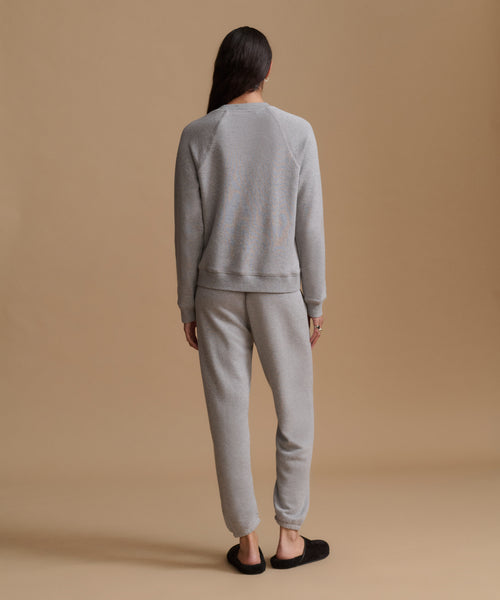 A woman viewed from behind wearing a light gray crewneck sweatshirt and matching jogger pants, paired with black slip-on slippers, against a solid warm beige studio background.