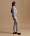 A woman in a side profile wearing a light gray knit lounge set consisting of a long-sleeve top and matching jogger pants, paired with black slip-on slippers against a solid beige studio background.