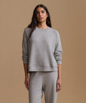 A woman modeling a light gray heather crewneck sweatshirt and matching sweatpants against a solid tan studio background.