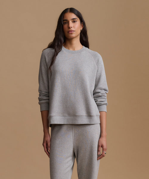 A woman modeling a light gray heather crewneck sweatshirt and matching sweatpants against a solid tan studio background.