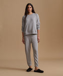 A woman modeling a light gray two-piece loungewear set consisting of a crewneck sweatshirt and matching jogger pants, paired with black slip-on slippers, against a solid beige studio background.