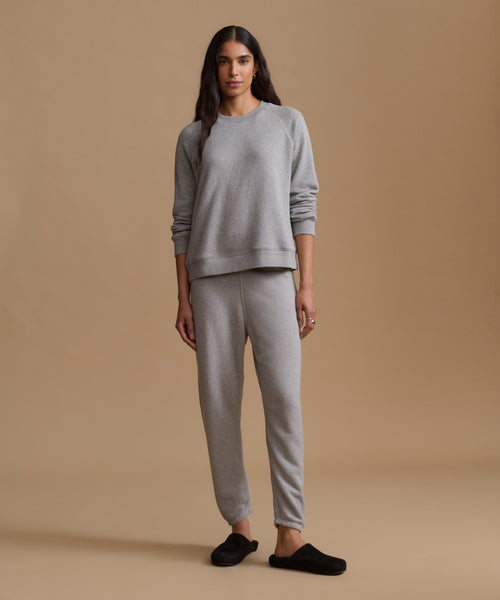 A woman modeling a light gray two-piece loungewear set consisting of a crewneck sweatshirt and matching jogger pants, paired with black slip-on slippers, against a solid beige studio background.