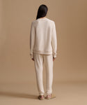 A model viewed from behind wearing a matching cream-colored knit lounge set consisting of a crewneck sweatshirt and jogger pants, paired with tan flats, against a solid beige studio backdrop.