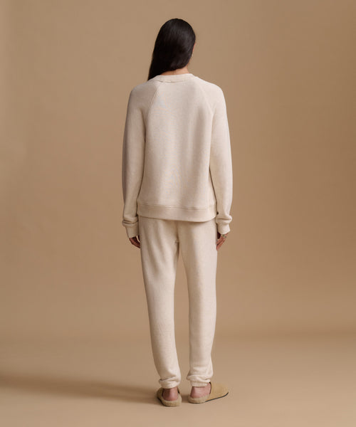 A model viewed from behind wearing a matching cream-colored knit lounge set consisting of a crewneck sweatshirt and jogger pants, paired with tan flats, against a solid beige studio backdrop.