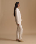 A woman stands in profile wearing a matching cream knit lounge set consisting of a long-sleeve top and wide-leg pants, paired with tan slip-on shoes, against a solid beige studio background.
