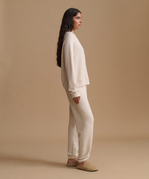 A woman stands in profile wearing a matching cream knit lounge set consisting of a long-sleeve top and wide-leg pants, paired with tan slip-on shoes, against a solid beige studio background.