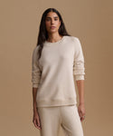 A woman modeling a matching cream-colored knit lounge set, featuring a crewneck sweatshirt and coordinating sweatpants, set against a solid warm beige studio background.