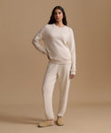 A woman modeling a matching cream knit lounge set consisting of a long-sleeve crewneck sweatshirt and relaxed-fit sweatpants, paired with tan slip-on slippers against a solid beige studio backdrop.