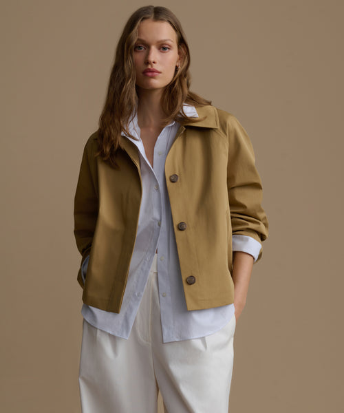 Woman wearing a mustard yellow jacket over a white shirt and beige pants on a brown background