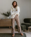 A smiling woman stands indoors in a white long-sleeve shirt, cream sweater over her shoulders, beige pants, and Shearling-Lined Moc Clogs. A wooden cabinet, books, and a green armchair are visible behind her.