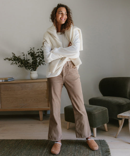 A smiling woman stands indoors in a white long-sleeve shirt, cream sweater over her shoulders, beige pants, and Shearling-Lined Moc Clogs. A wooden cabinet, books, and a green armchair are visible behind her.