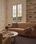 A cozy living room with a beige sofa, the Sheridan Pillow, and a brown throw blanket. A round wooden coffee table holding flowers and books sits on a dark rug, while large windows fill the space with natural light.