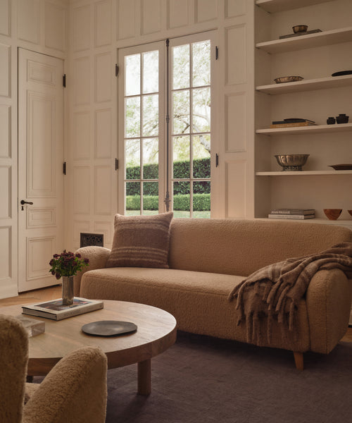 A cozy living room with a beige sofa, the Sheridan Pillow, and a brown throw blanket. A round wooden coffee table holding flowers and books sits on a dark rug, while large windows fill the space with natural light.