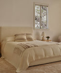 A cozy bedroom features a cream bed with two striped pillows and the soft Sheridan Throw. Wooden nightstands hold a lamp and a small plant, while the window frames tree branches outside.