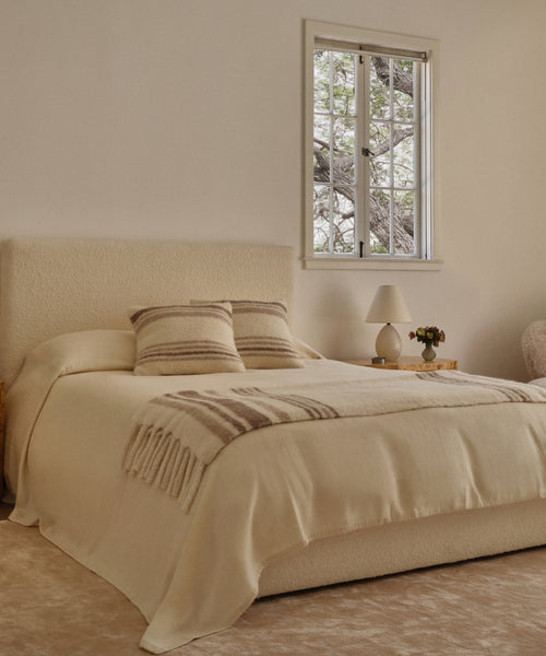 A cozy bedroom features a cream bed with two striped pillows and the soft Sheridan Throw. Wooden nightstands hold a lamp and a small plant, while the window frames tree branches outside.