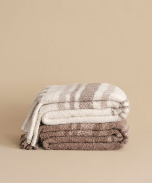 Two folded Sheridan Throws rest on a beige surface: the top features cream and wide tan stripes, while the bottom is solid light brown. Both look soft and plush—perfect for pairing with a Sheridan pillow.