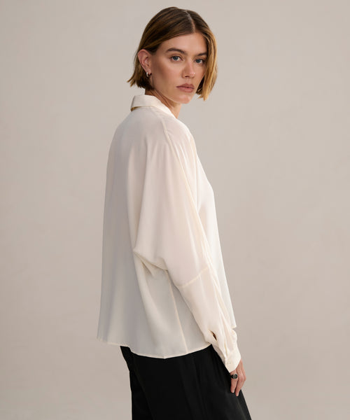 A person with short, light brown hair stands sideways, looking over their shoulder in a loose Silk Sloane Shirt and black pants against a plain, light background.