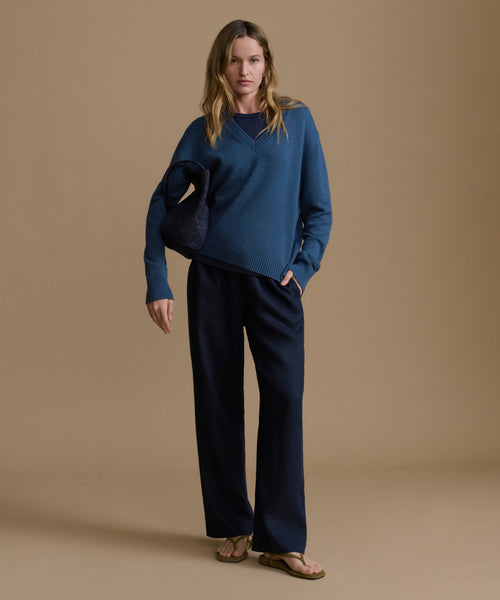 Woman wearing a navy v-neck sweater with blue pants against a brown wall. 