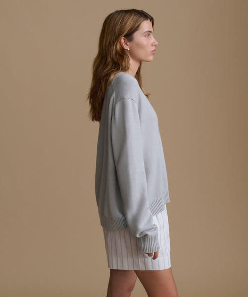Woman wearing a light blue v-neck sweater with blue and white stripe shorts against a brown wall. 