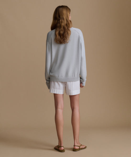 Woman wearing a light blue sweater with blue and white stripe shorts against a brown wall. 