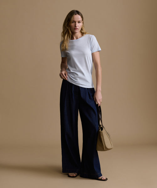 Woman wearing a baby blue t-shirt and navy pants holding a beige handbag on a brown background