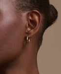 Close-up of a person’s ear adorned with Jenni Kayne Small Eloise Hoops in 14k gold. Hair is pulled back, highlighting smooth skin and the elegant, timeless touch of this heirloom jewelry against a plain beige background.