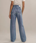 A person stands on a light surface facing away, wearing The Standard: cream short-sleeve shirt and higher mid-rise, straight-leg jeans with visible back pockets and seams.