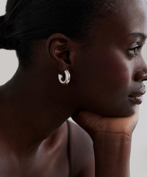 A woman with dark skin and a smooth complexion, in a sleeveless top, rests her chin on her hand and gazes thoughtfully to the right, showing off sculptural Small Donut Hoops in sterling silver.