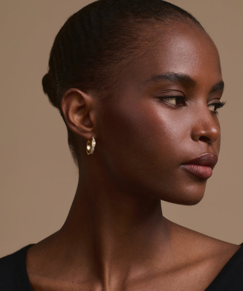 A woman with smooth dark skin and braided hair pulled back looks to the right. She is wearing Stevie Grey Diamond Hoops and a black top against a neutral beige background.
