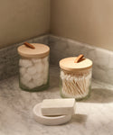 Two Stowe Eco Glass Canisters with wooden lids, one holding cotton balls and the other cotton swabs, rest on a marble countertop beside a white soap bar, bringing sustainable storage to a softly lit bathroom corner.