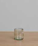 The Straight Rock Glass, handblown from recycled glass with a light green tint, rests on a pale wooden surface against a plain light gray background—a perfect sustainable glassware choice.