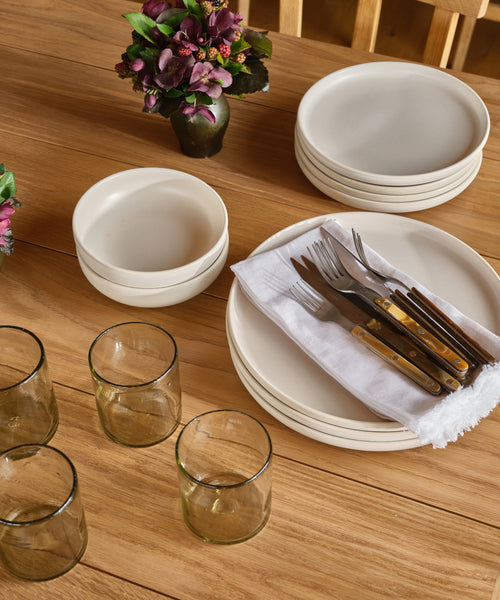 A wooden table set with stacked white plates and bowls, a cloth napkin with forks and knives, small vases of purple flowers, and handblown recycled Straight Rock Glasses.