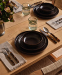 A dining table set with ceramic dinnerware and recycled glass cups with vases filled with florals. 