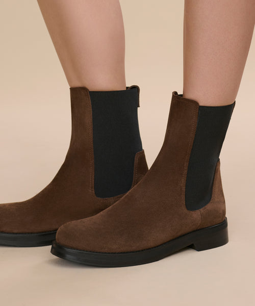 A close-up of a person wearing the Suede Alden Boot—brown suede Chelsea boots with black elastic side panels and a weather-friendly rubber sole—standing on a light beige surface.