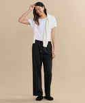 A woman stands against a beige background, smiling. She wears a white T-shirt, black pants, and Suede Ashby Clogs with a shearling-lined footbed, plus a cream sweater draped over her shoulders. One hand touches her head.