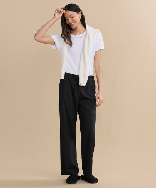 A woman stands against a beige background, smiling. She wears a white T-shirt, black pants, and Suede Ashby Clogs with a shearling-lined footbed, plus a cream sweater draped over her shoulders. One hand touches her head.