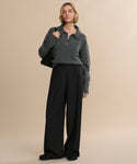 A person stands against a beige background wearing a gray pullover, loose black trousers, black shoes, and a black bag, with short wavy hair completing the look—finished with classic Suede Ashby Clog footwear.