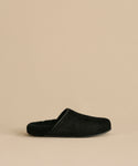 The Suede Ashby Clog, a black slip-on with a fuzzy shearling-lined footbed and closed toe, is shown against a beige background.