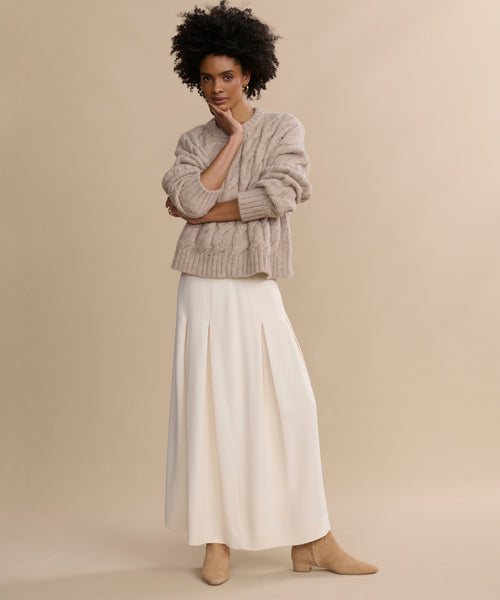 A woman with curly hair stands against a beige background, wearing a chunky beige sweater, pleated long white skirt, and Suede Carmine Boots in taupe. She rests her chin on her hand and gazes thoughtfully at the camera.