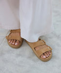 A person wearing wide-legged light pants and the Suede Hutton Slide Sandal with signature push-button details stands on a light gray textured surface, their neatly manicured toes highlighting this ideal warm-weather shoe.